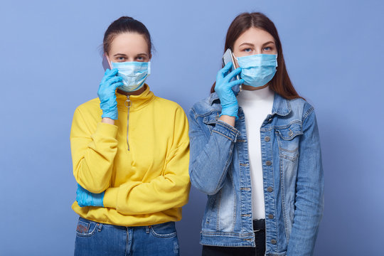 Picture of two beautiful young friends holding smartphones in hands, talking over phone, looking directly at camera, being in masks and gloves, communicating in distance. Coronavirus concept.