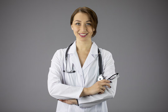 Portrait of an attractive sexy doctor woman in a white medical coat with a stethoscope on her neck. Hands are folded on the chest. Red painted lips, a snow-white smile. Grey background. Studio shot