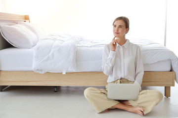 young attractive caucasian woman wearing white casual clothes sitting and working on computer while thinking something in bedroom (internet of thing and life style concept)