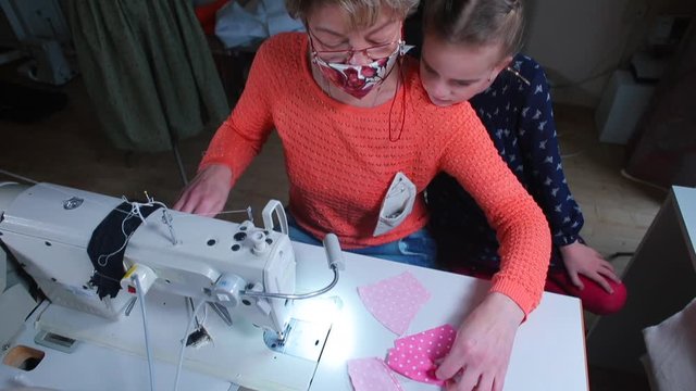  Home education COVID-19. Grandmother and granddaughter sew a face mask on a sewing machine. Useful things in self-isolation. Joining the edges of the fabric with thread.