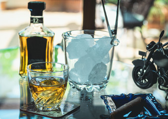Glass of whiskey and cigar next to a bottle and an ice bucket on a glass table