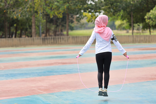 Rear view athletic young asian muslim woman in sportswear standing and  skipping rope outdoor for morning exercise. Active girl work out on biceps exercise with green tree background. Sport concept