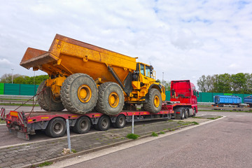 Big truck with a low platform trailer carrying a tipper truck on a public parking area of a truck stop.