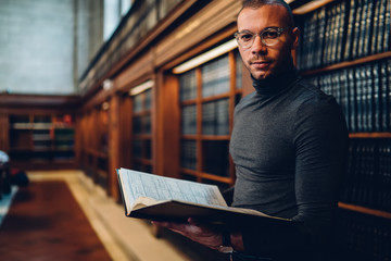 Half length portrait of intellectual professor of history in bifocal eyewear looking at camera while holding literature book in hand for making research of information for new science publication