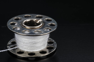 Close up of a metal sewing machine bobbin with white thread on a black background.