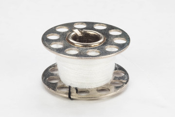 Close up of a metal sewing machine bobbin with white thread on a white background.