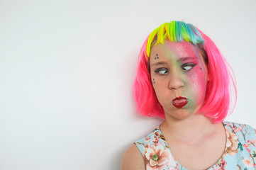 Teenage girl with a multi coloured wig and heavy make-up against a white wall.