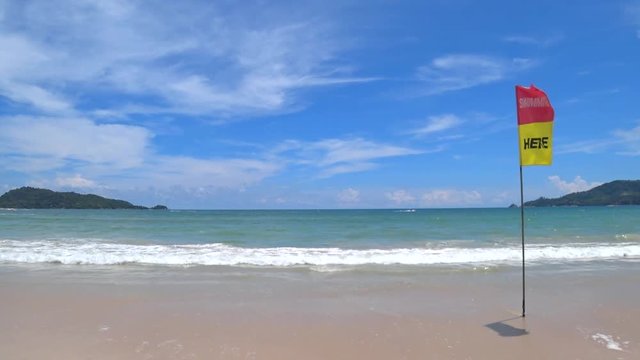White foam wave running to the powdery sand with red and yellow "swimming here" flag, nobody swim in the ocean, on a bright and sunny day at Patong beach, Phuket, Thailand.