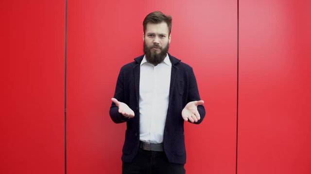 Young brunette bearded man is gesturing and talking something to the camera against red background. Guy in black jacket and white shirt is explaining or giving advice near red wall.