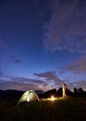 Male tourist spending time in the mountains in the evening with illuminated tent and a campfire...
