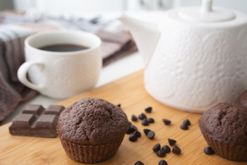 Chocolate muffins, chocolate chips, chocolate bar, a cup of coffee, teapot on a wooden surface with a warm scarf, Christmas lights bokeh and holly in the background