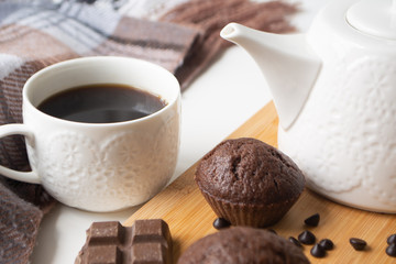 Chocolate muffins, chocolate chips, chocolate bar, a cup of coffee, teapot on a wooden surface with a warm scarf, Christmas lights bokeh and holly in the background