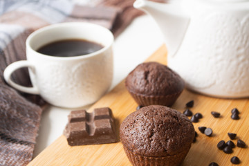 Chocolate muffins, chocolate chips, chocolate bar, a cup of coffee, teapot on a wooden surface with a warm scarf, Christmas lights bokeh and holly in the background
