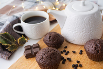 Chocolate muffins, chocolate chips, chocolate bar, a cup of coffee, teapot on a wooden surface with a warm scarf, Christmas lights bokeh and holly in the background