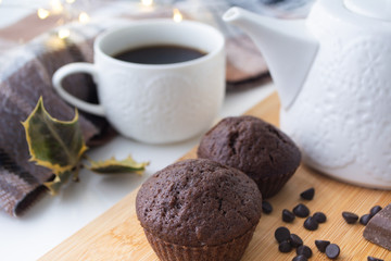 Chocolate muffins, chocolate chips, chocolate bar, a cup of coffee, teapot on a wooden surface with a warm scarf, Christmas lights bokeh and holly in the background