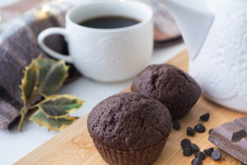 Chocolate muffins, chocolate chips, chocolate bar, a cup of coffee, teapot on a wooden surface with a warm scarf, Christmas lights bokeh and holly in the background
