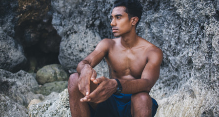 Pensive Hispanic man in shorts sitting near cave