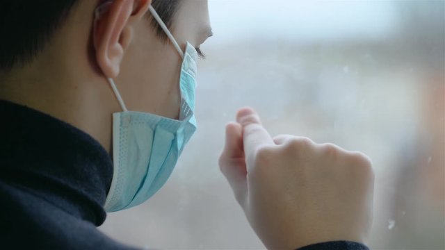 Sad boy in medical mask draws finger on window out of boredom during world corona virus pandemic. Stop COVID-19 infection