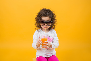 Little girl in a glass drink orange juice on a yellow background. Children's emotion, humor, space for text.
