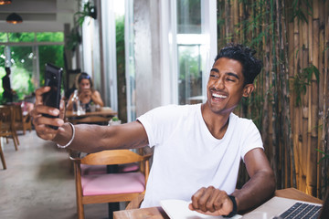 Smiling adult ethnic man taking selfie with smartphone in cafe