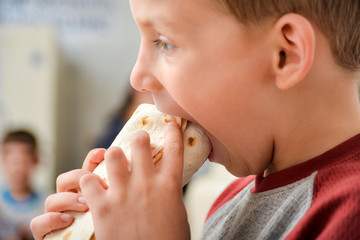 The boy holds shawarma in his hands, the child bites off meat wrapped in pita bread.