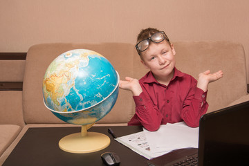 a boy in glasses and a red shirt is sitting at a black table looking at a geographical globe and doing homework, a child in quarantine on distance learning. he has ptosis in his left eye.
