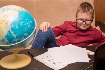 a boy in glasses and a red shirt is sitting at a black table looking at a geographical globe and doing homework, a child in quarantine on distance learning.