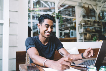 Joyful ethnic student taking notes in notebook while surfing laptop in cafe
