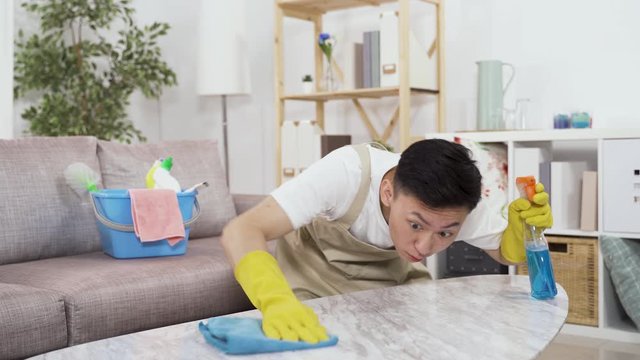 chinese male neat freak in apron and rubber gloves wiping table in parlor is trying to remove stain. asian guy bending and holding cleanser spray is sanitizing home to prevent virus.