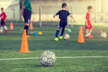 Fototapeta premium Old soccer ball on green artificial turf with blurry soccer team training. Blurry kid soccer player jogging between marker cones