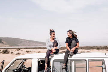 Two happyyoung girls laughing and having fun while sitting on top of the van while traveling. These two girls love adventure, roadtrips and become wild to pursue happiness. Travel Concept. Copyspace 