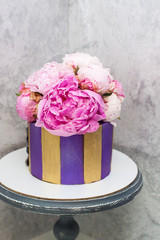 Modern, elegant violet and golden cake decorated with fresh peony flowers. Grey background