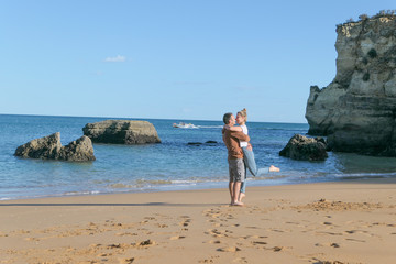 A couple near the blue sea and big rude rocs. Yellow sand on the beach.