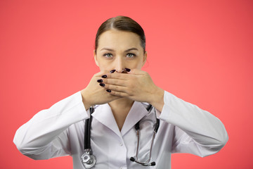 Beautiful young sexy doctor in white medical coat and stethoscope closes mouth by hands, seriously looking at camera on isolated red background. Secret, silence concept
