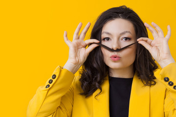 Stylish young woman in a yellow jacket on a yellow background, making a mustache out of her hair.