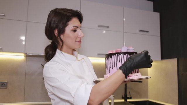 Woman baker is decorating a birthday cake with macarons at the top. The cake is decorated with chocolate topping and looks mouth-watering.