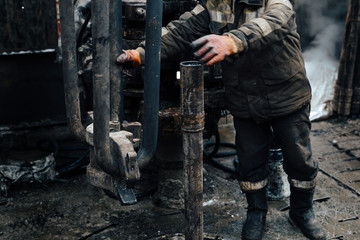 Work driller in red uniform, in helmet and goggles. He uses a hydraulic wrench to screw drill pipes to lower them into an oil well and continue drilling it. The concept of a working person.
