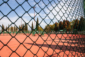 Empty tennis court with orange coating behind metal fence net, cypresses and another autumn trees behind, blue sky, day light 
