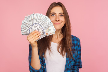 Portrait of happy lucky girl in checkered shirt holding lot of money and smiling to camera, showing dollar bills in hand, enjoying financial success. indoor studio shot isolated on pink background