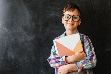 Cute boy, a schoolboy in glasses stands on the background of a black school board, holds a book in his hands, smiles.school education.place for text