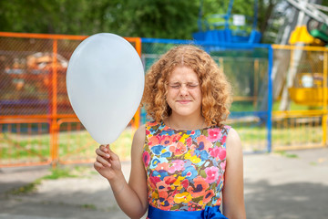 The red-haired girl in a colored dress holds a white ball in her hand and squeezed her eyes shut.