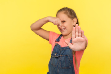 Awful smell. Little girl with braid in denim overalls holding breath, pinching her nose and showing stop gesture, expressing disgust to stink, fart. indoor studio shot isolated on yellow background