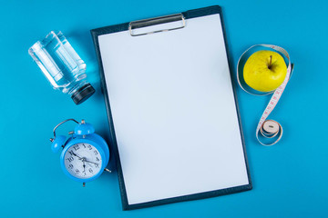 The diet plan. White sheet on the tablet among the bottles with water, clock and apples with...