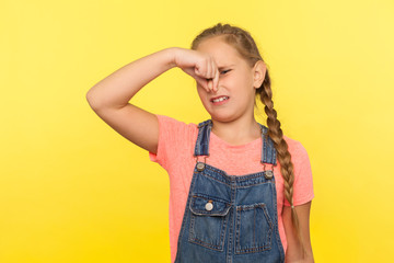 Awful smell. Portrait of little girl with braid in denim overalls holding breath, pinching her nose...