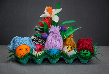 Orange, red, blue, yellow and green Easter eggs in blue, black and white, lilac, terracotta and red knitted hats in a green basket on a gray background with red, white and purple flowers made of paper