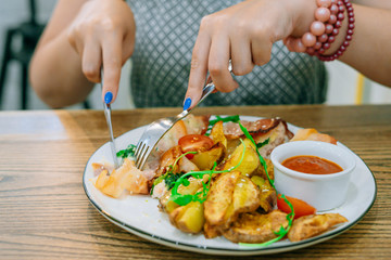 Female hands with knife and fork cut chicken in bacon. Chicken, potatoes and sauce. Female hands with manicure and bracelet
