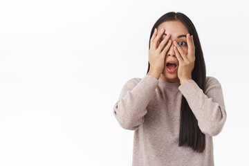 Scared embarrassed cute asian girl hiding face, hold hands above eyes and peeking at camera, frightened or alarmed, watching horror movie alone dark, afraid clowns, standing white background shook