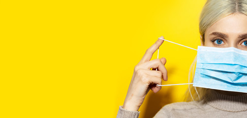 Half face portrait of young blonde girl with blue eyes putting on medical flu mask on face, on yellow panoramic background with copy space.