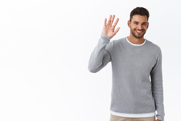 Friendly modest handsome bearded man in grey sweater saying hi with hesitation lifting one palm,...