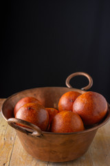 Aerial view of copper bowl with blood oranges on rustic wooden table, with black background, vertical